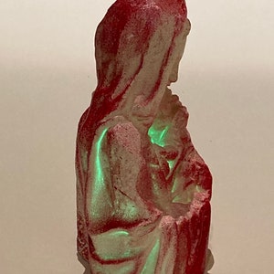 May include: A vintage Christmas ornament depicting the Virgin Mary holding baby Jesus. The ornament is made of plaster and painted in shades of red and white. The ornament glows green in the dark.