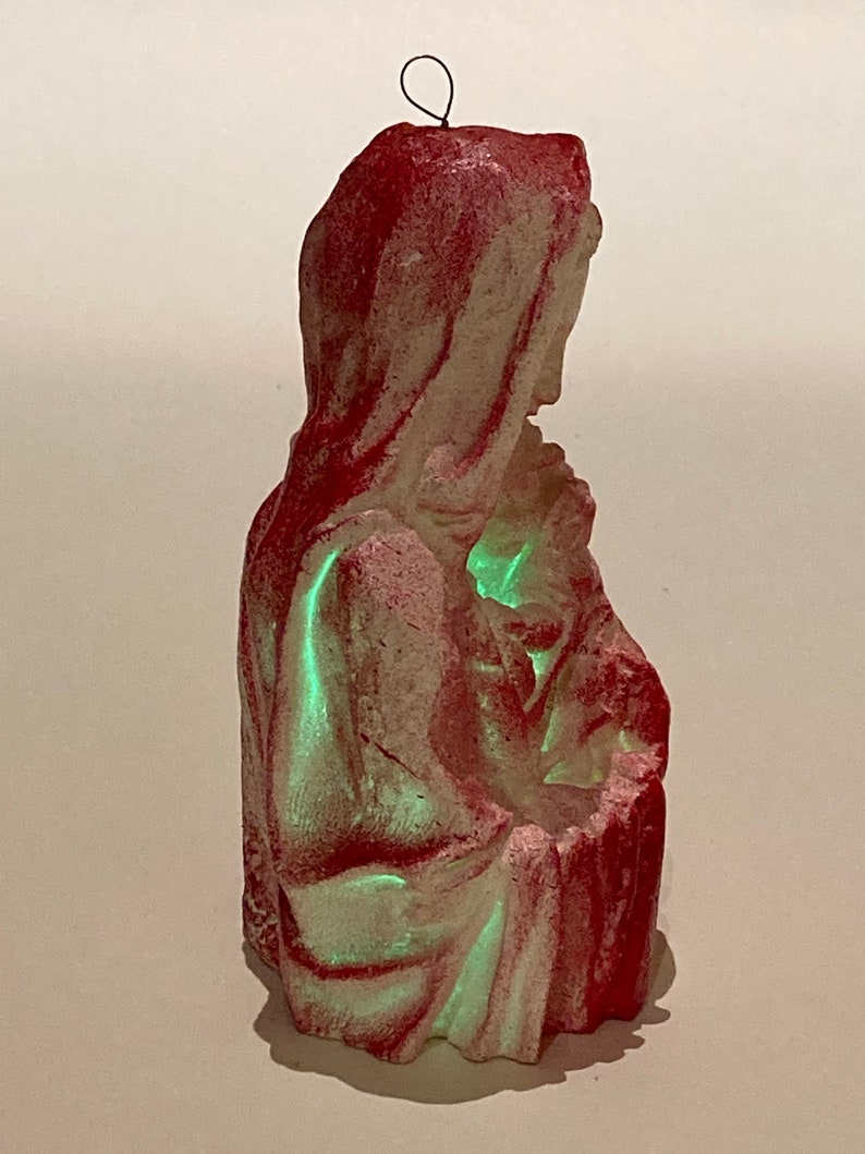 May include: A vintage Christmas ornament depicting the Virgin Mary holding baby Jesus. The ornament is made of plaster and painted in shades of red and white. The ornament glows green in the dark.