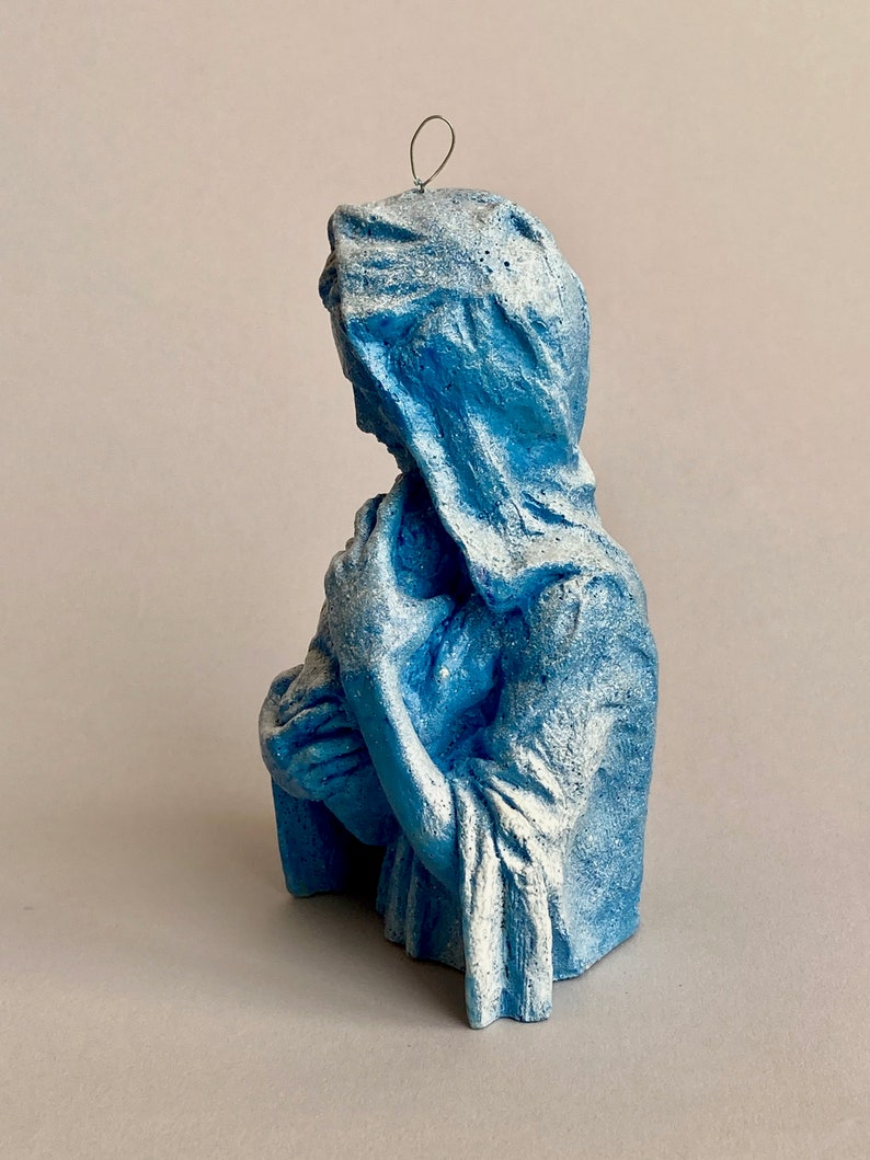 May include: A blue ceramic figurine of the Virgin Mary holding a baby Jesus. The figurine is painted in a light blue color with white accents.