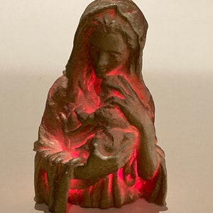 May include: A beige ceramic figurine of the Virgin Mary holding a baby Jesus. The figurine is illuminated from within with a red light.