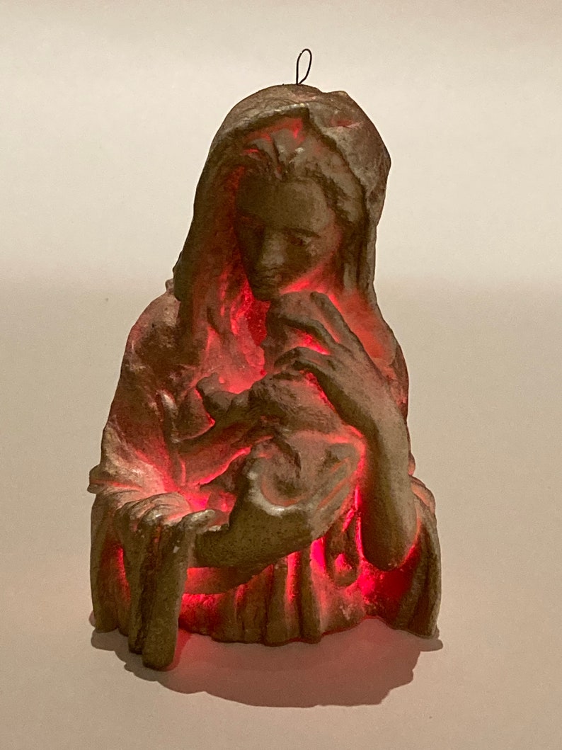 May include: A beige ceramic figurine of the Virgin Mary holding a baby Jesus. The figurine is illuminated from within with a red light.