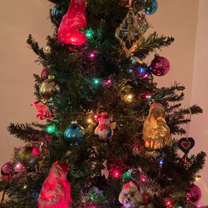 May include: A decorated Christmas tree with colorful lights, ornaments, and figurines. The ornaments include a snowman, a white dog, a red angel, a gold religious figure, and a heart-shaped ornament.