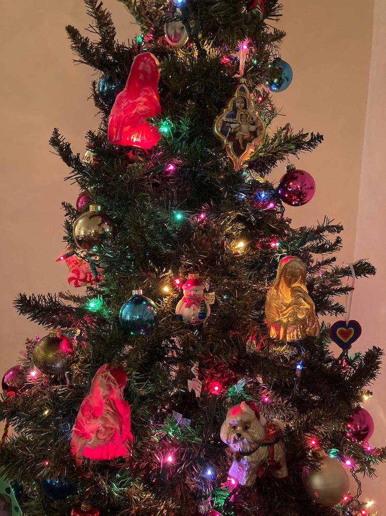 May include: A decorated Christmas tree with colorful lights, ornaments, and figurines. The ornaments include a snowman, a white dog, a red angel, a gold religious figure, and a heart-shaped ornament.