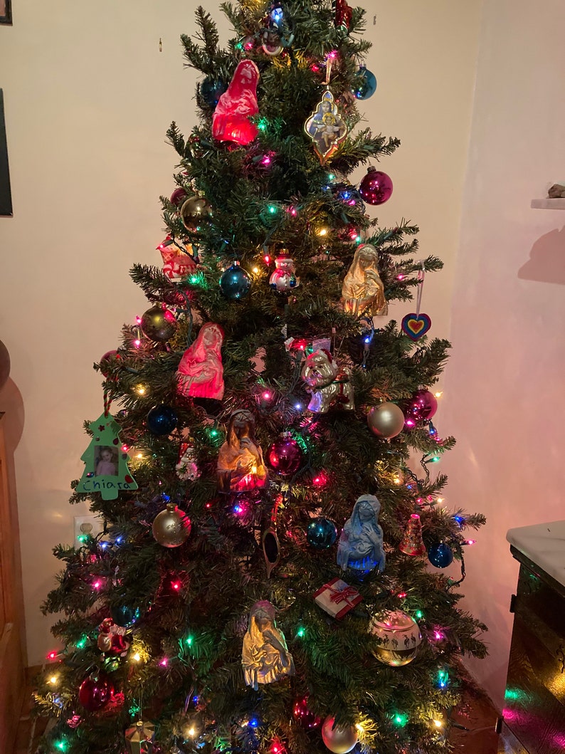 May include: A decorated Christmas tree with colorful lights and ornaments, including a blue, red, and gold figurine of the Virgin Mary, a wooden ornament with a child's photo and the name "Chiara", and a heart-shaped ornament.