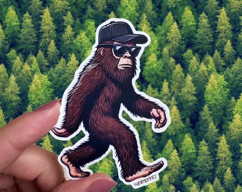 May include: A brown Bigfoot sticker with a black baseball cap and sunglasses. The sticker is on a green background of pine trees.