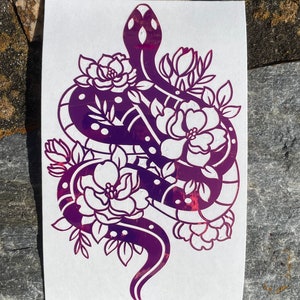 May include: A purple vinyl sticker of a snake wrapped around flowers. The snake is outlined in black and has a dotted pattern. The flowers are white and have a detailed design.