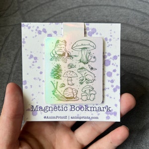 May include: A holographic magnetic bookmark with a design of a frog, snail, mushrooms, and a pine tree. The bookmark has the text "Magnetic Bookmark" and "@AnimPrintZ | animprintz.com".