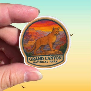 May include: A round sticker featuring a cougar standing on a rock with the Grand Canyon in the background. The sticker has a sunset sky and the text "GRAND CANYON NATIONAL PARK". The sticker is held by a hand.