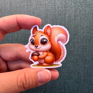 May include: A cartoon sticker of a cute, brown squirrel holding an acorn. The squirrel has big, round eyes and a bushy tail.