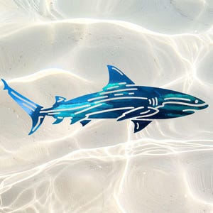 Puede incluir: Una pieza de arte mural de metal con forma de tibur&oacute;n, que presenta un acabado azul profundo con recortes lineales en azul claro y blanco que crean una sensaci&oacute;n de movimiento y textura en el cuerpo, las aletas y la cola del animal.