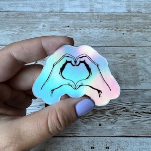 May include: A holographic sticker with a black outline of two hands forming a heart shape.
