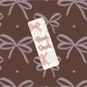 May include: A rectangular bookmark with a light pink background, featuring a skeleton hand with a pink bow, the words "Book Snob", and floral accents. The background is a pattern of brown bows.