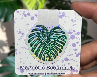 May include: A magnetic bookmark featuring a dark green Monstera leaf illustration with a holographic glitter finish. The bookmark is attached to a white backing card with purple paint splatter accents and the text Magnetic Bookmark @AnimPrintZ | animprintz.com.