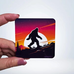 May include: A sticker featuring a silhouette of Bigfoot walking across a mountain range at sunset. The background is a gradient of orange, pink, and purple with a full moon in the sky.