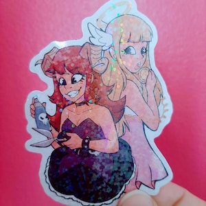 May include: A holographic sticker depicting two cartoon characters: a devilish figure with horns and a dark dress holding scissors and a skull-adorned spray can, and an angelic figure with wings and a halo. Set against a pink backdrop.