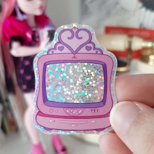 May include: A pink and purple glitter television sticker with a crown on top. The television screen is filled with iridescent glitter.