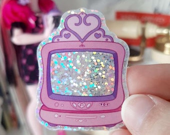 May include: A pink and purple glitter television sticker with a crown on top. The television screen is filled with iridescent glitter.