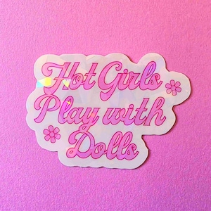 May include: A die-cut sticker with a holographic finish featuring the pink text Hot Girls Play with Dolls in a cursive font. The design includes two small pink flower icons, one on the left and one on the right, against a reflective background.