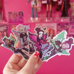 May include: Three holographic stickers featuring fairy characters with wings. The fairies are wearing colorful outfits and have different hairstyles. The stickers are on a pink background.