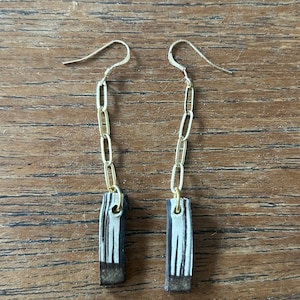 Könnte beinhalten: Ein Paar Ohrhänger mit goldfarbenen Metallkettengliedern, die an rechteckigen Anhängern befestigt sind. Die Anhänger haben ein vertikales Streifenmuster in Schwarz, Weiß und Braun mit einer soliden dunkelbraunen Basis am unteren Ende.