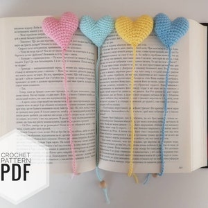 May include: Four crochet heart bookmarks in pink, light blue, yellow, and blue, each attached to a string and placed in a book. The text "CROCHET PATTERN PDF" is visible in the lower left corner.