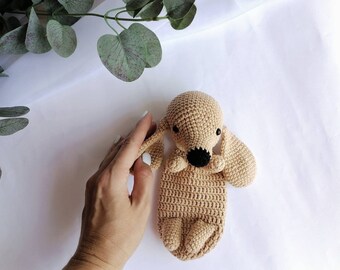 Op de afbeelding: Een gehaakt knuffeldier in de vorm van een hond in een lichtbruine kleur met zwarte ogen en neus. Het speelgoed ligt op een wit oppervlak.