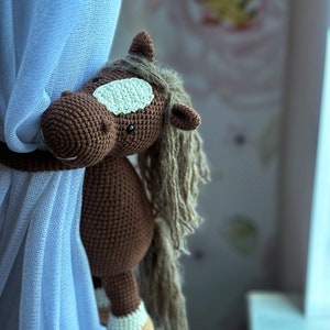 May include: A brown crocheted horse toy with a white patch on its face and a long brown mane. The horse is hanging from a blue curtain.