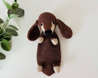 Op de afbeelding: Een bruin gehaakt knuffeldier in de vorm van een teckel met witte accenten op de poten en oren. Het speelgoed ligt op zijn rug met zijn poten in de lucht.