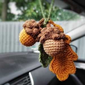 May include: A crocheted acorn charm with a brown and tan color scheme, hanging from a car's rearview mirror. The acorn is attached to a crocheted leaf with a brown and orange color scheme.