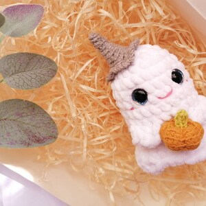 May include: White crocheted ghost holding a crocheted orange pumpkin. The ghost has black eyes and a pink smile. The ghost is sitting in a bed of shredded paper.