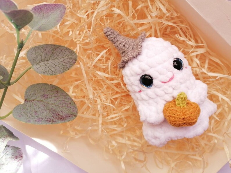 May include: White crocheted ghost holding a crocheted orange pumpkin. The ghost has black eyes and a pink smile. The ghost is sitting in a bed of shredded paper.