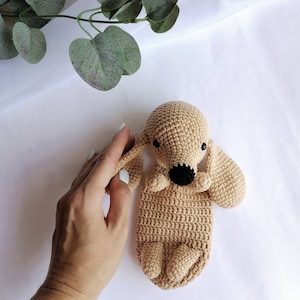 Op de afbeelding: Een gehaakt knuffeldier in de vorm van een hond in een lichtbruine kleur met zwarte ogen en een zwarte neus. Het speelgoed ligt op een wit oppervlak.