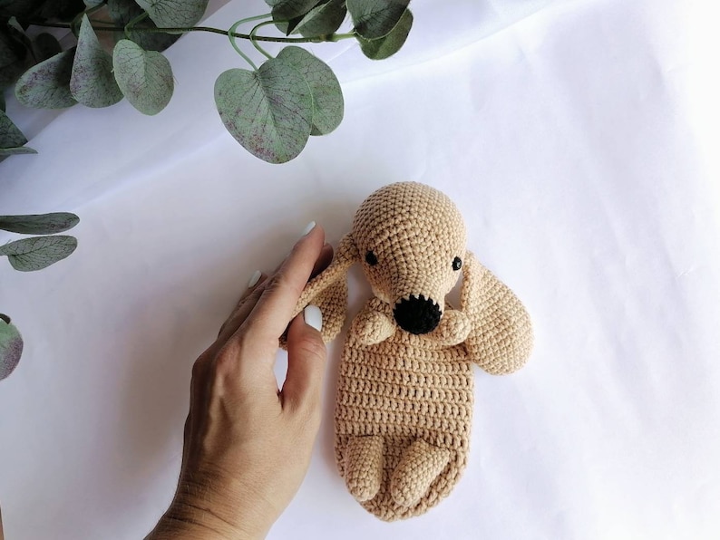 Op de afbeelding: Een gehaakt knuffeldier in de vorm van een hond in een lichtbruine kleur met zwarte ogen en een zwarte neus. Het speelgoed ligt op een wit oppervlak.