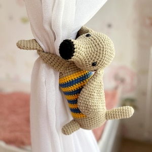 May include: A crocheted dog-shaped curtain tieback in beige with a black nose and blue and yellow striped shirt. The dog is holding a sheer white curtain. The background is a soft pink and white floral pattern.