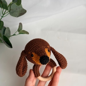 May include: A crocheted dog-shaped baby rattle with brown and tan yarn. The dog has long floppy ears and a black nose. The rattle features a natural wooden ring for easy gripping. The toy is designed for infants and toddlers.