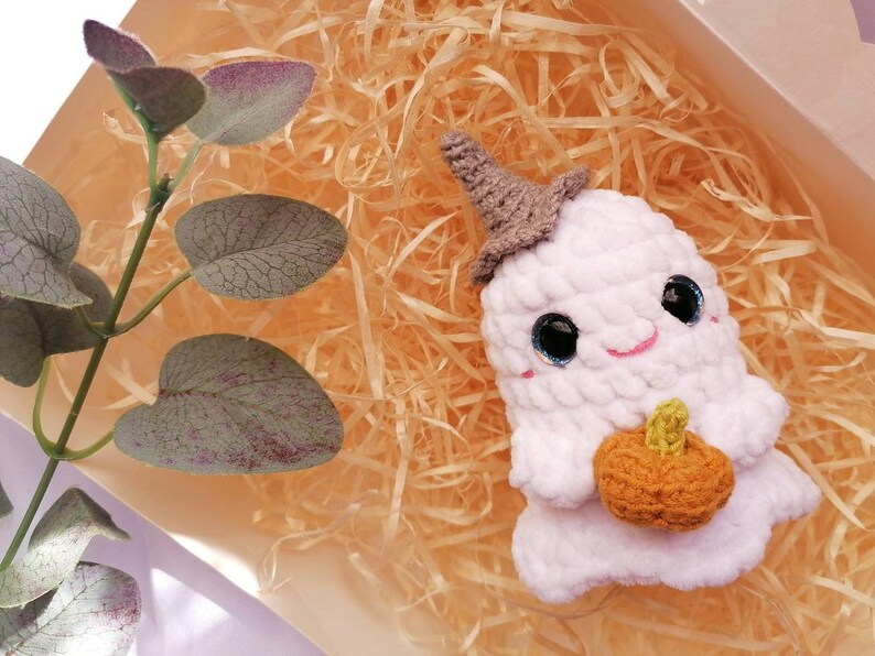 May include: A white crocheted ghost holding a small orange crocheted pumpkin. The ghost is wearing a gray crocheted witch hat. The ghost is sitting in a bed of shredded paper.