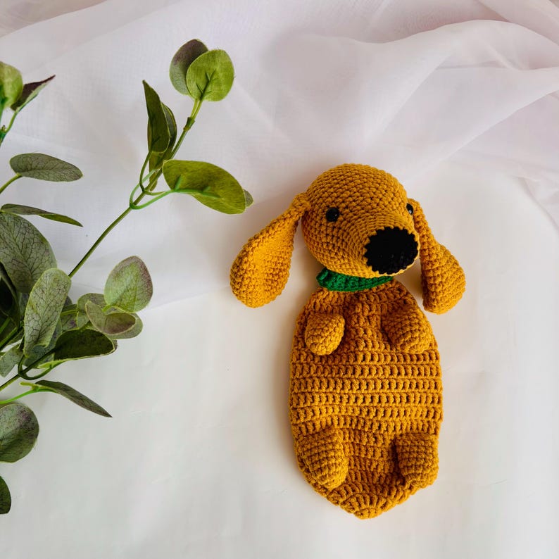 Op de afbeelding: Een gehaakt knuffeldier in de vorm van een hond in mosterdgele kleur met een groene halsband. De hond ligt op zijn buik met zijn poten onder zijn lichaam.