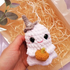 May include: A white crocheted ghost holding a small orange crocheted pumpkin. The ghost has a black hat and black eyes with pink cheeks.