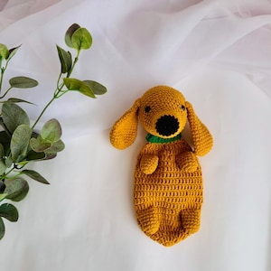 Op de afbeelding: Een gehaakt knuffeldier in de vorm van een hond in mosterdgele kleur met een groene halsband en een zwarte neus. Het speelgoed ligt op een wit oppervlak.