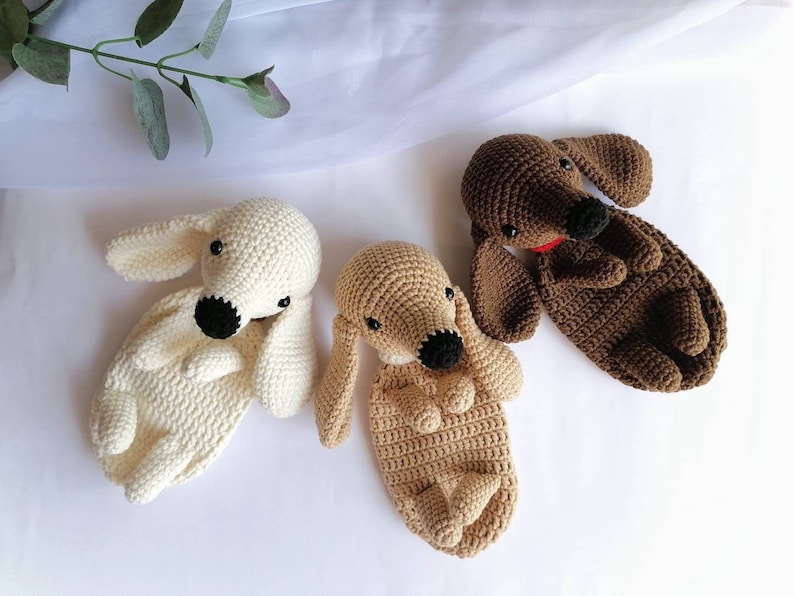 Op de afbeelding: Drie gehaakte teckel knuffels in verschillende kleuren: wit, bruin en beige. De knuffels liggen op een wit oppervlak.