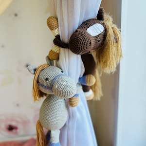 May include: Two crocheted horse toys hanging on a white curtain. The larger horse is brown with a white face and a long mane. The smaller horse is gray with a blue bridle and a brown mane.