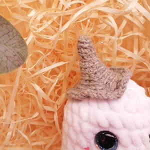 May include: A pink crocheted stuffed animal with a brown crocheted hat. The animal has large black eyes and a small pink nose.