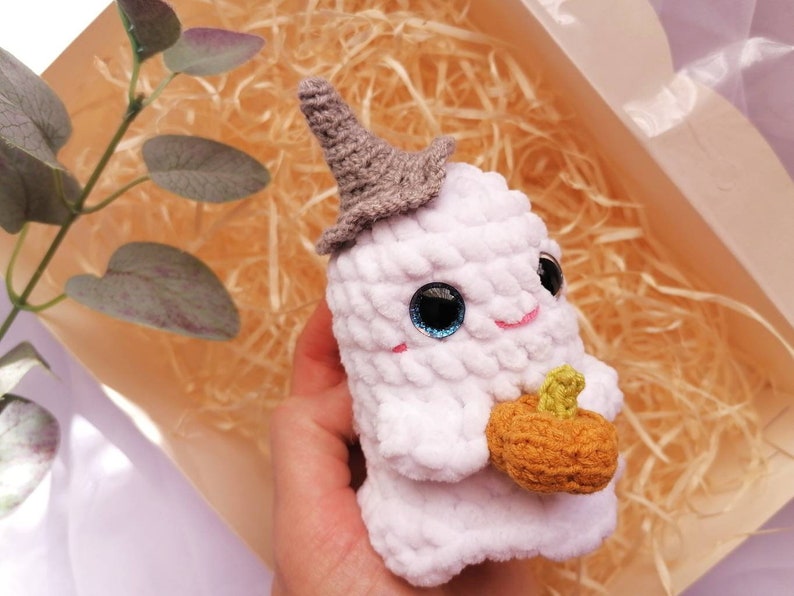 May include: White crocheted ghost holding a small orange pumpkin. The ghost has a gray pointed hat and blue eyes.