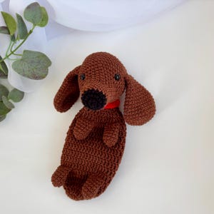 Op de afbeelding: Een bruin gehaakt knuffeldier in de vorm van een teckel met zwarte ogen en een rode halsband.