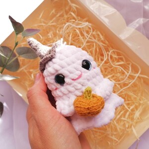 May include: A white crocheted ghost holding a small orange crocheted pumpkin. The ghost has a gray pointed hat and black eyes. The ghost is sitting in a box filled with shredded paper.