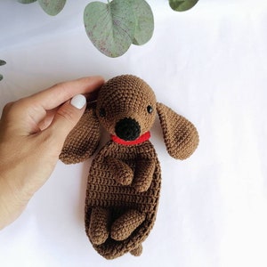 Op de afbeelding: Een bruin gehaakt knuffeldier in de vorm van een teckel met een rode halsband. Het speelgoed ligt op een wit oppervlak.