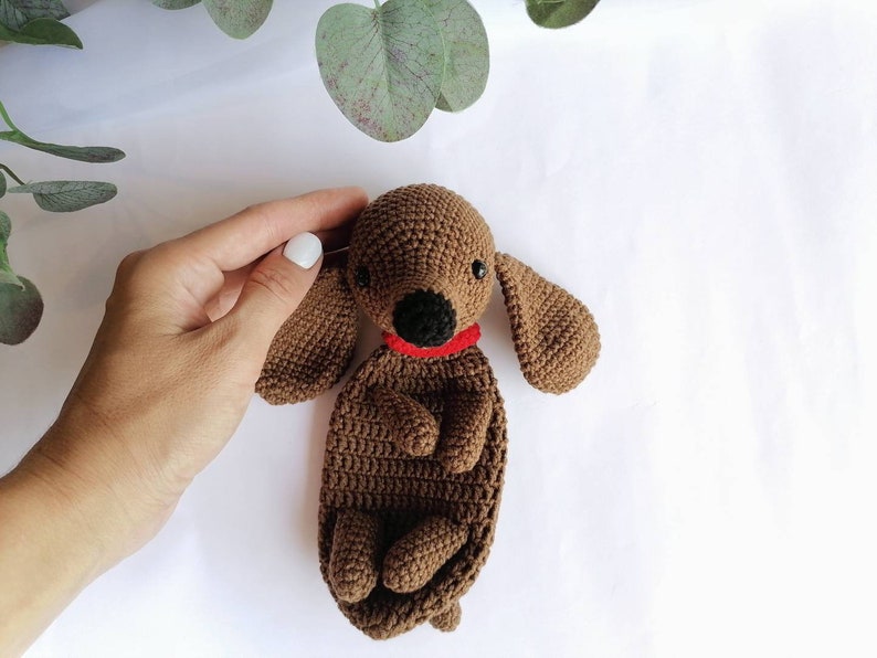Op de afbeelding: Een bruin gehaakt knuffeldier in de vorm van een teckel met een rode halsband. Het speelgoed ligt op een wit oppervlak.