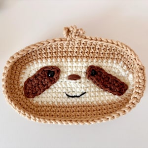 May include: A crocheted sloth-shaped basket with a brown face, white fur, and brown eyes. The basket is made of beige yarn and has a brown crocheted border.