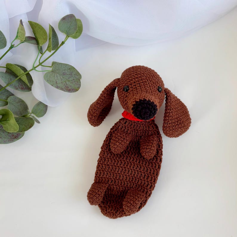 Op de afbeelding: Een bruin gehaakt knuffeldier in de vorm van een teckel met een rode halsband. Het speelgoed ligt op zijn rug met zijn poten in de lucht.
