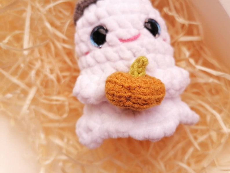 May include: A white crocheted ghost holding a small orange crocheted pumpkin. The ghost has black eyes and a pink mouth.